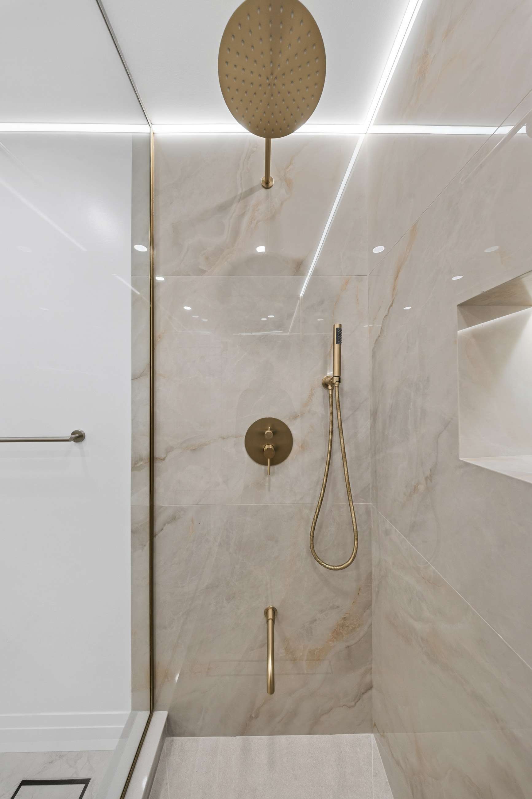 Brushed brass rain head and hand shower against stone, beside the illuminated niche.