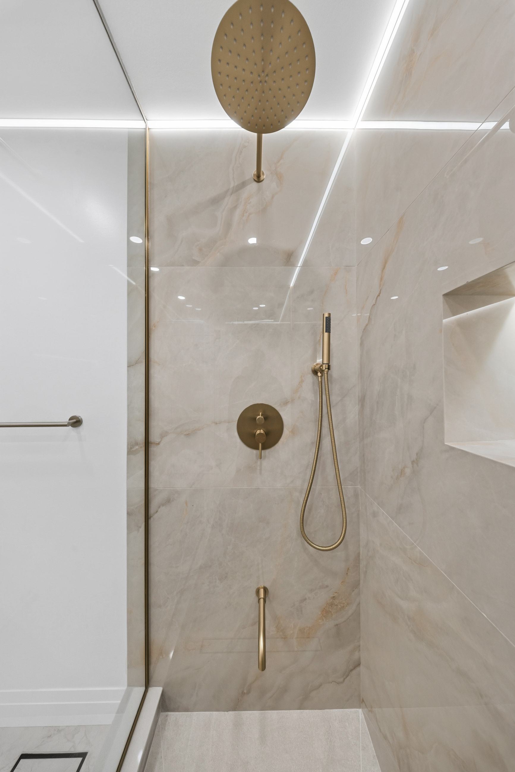 Brushed brass rain head and hand shower against stone, beside the illuminated niche.