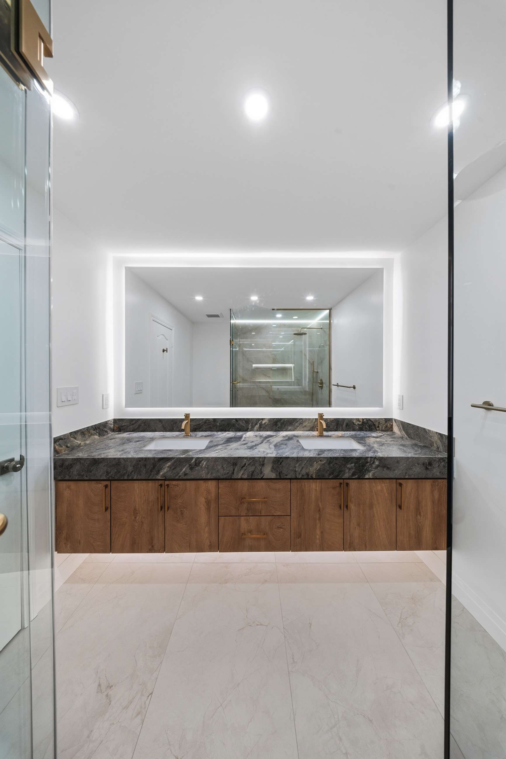 The vanity square on: twin brushed brass faucets on a stone counter under a backlit mirror.