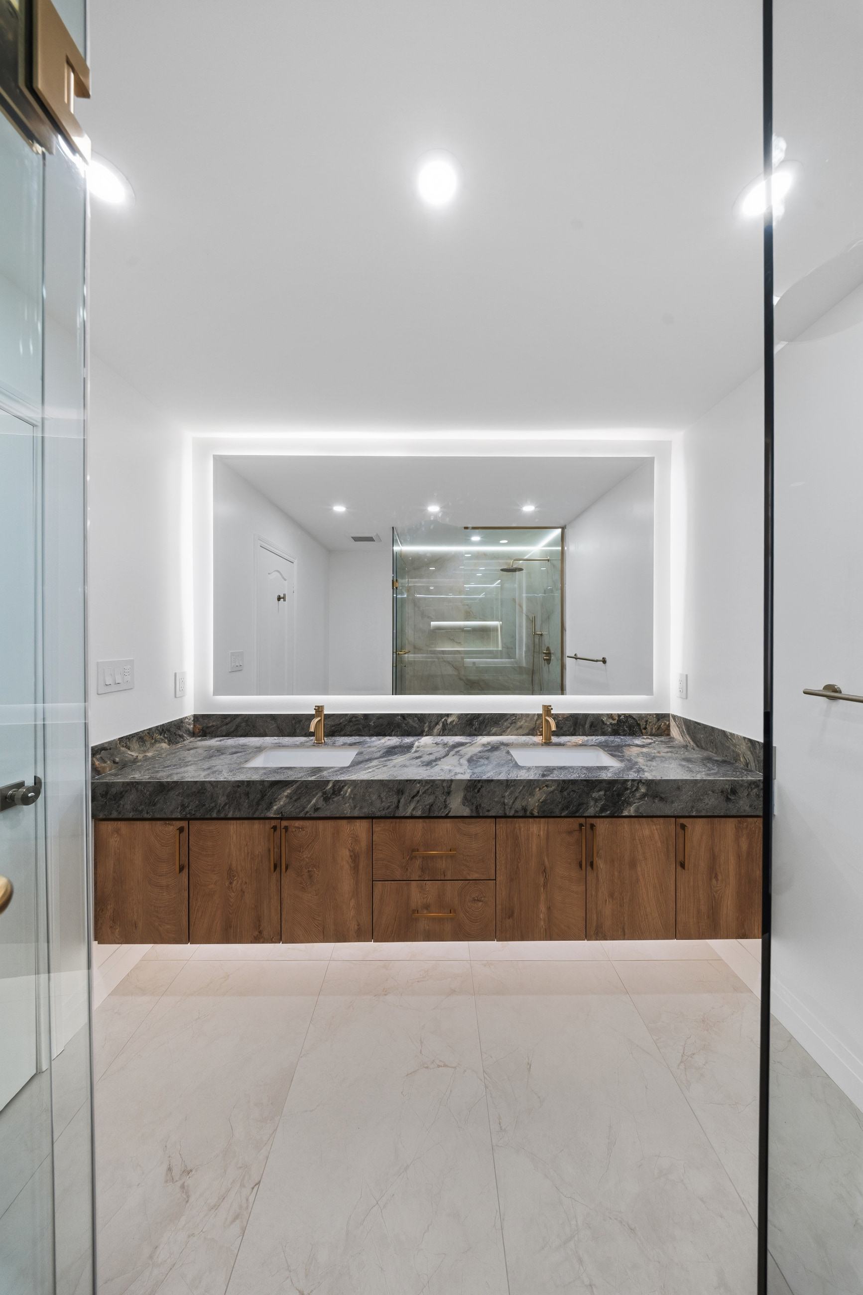 The vanity square on: twin brushed brass faucets on a stone counter under a backlit mirror.
