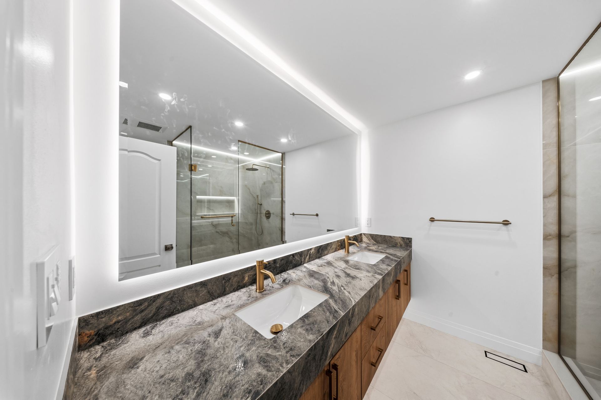 Primary ensuite renovation by TruBilt: a wide stone vanity with brushed brass faucets, walnut cabinetry and a backlit mirror, with a framed glass shower beyond.