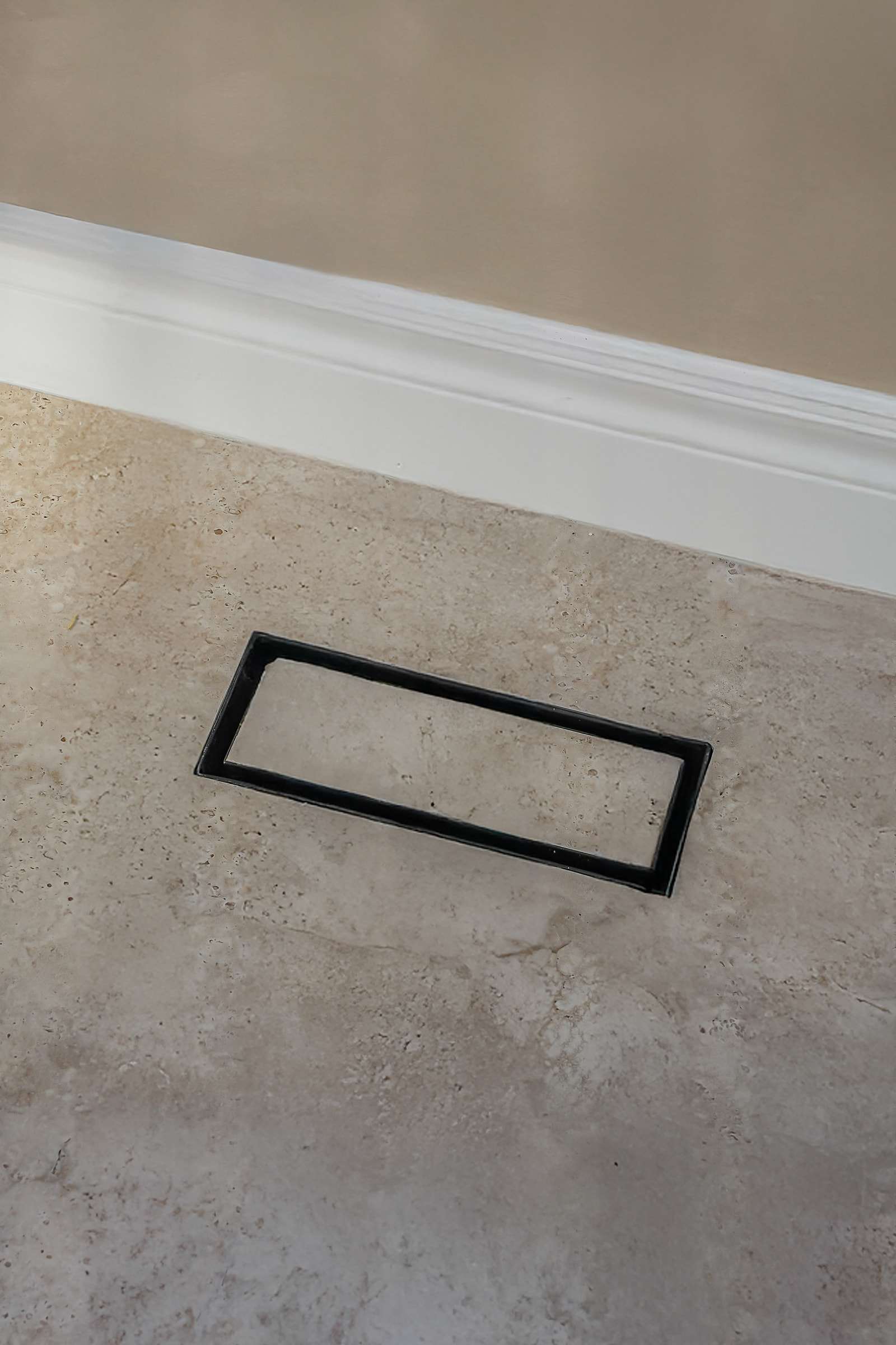 A rectangular floor vent near the baseboard, filled with the floor tile so the surface runs through it.