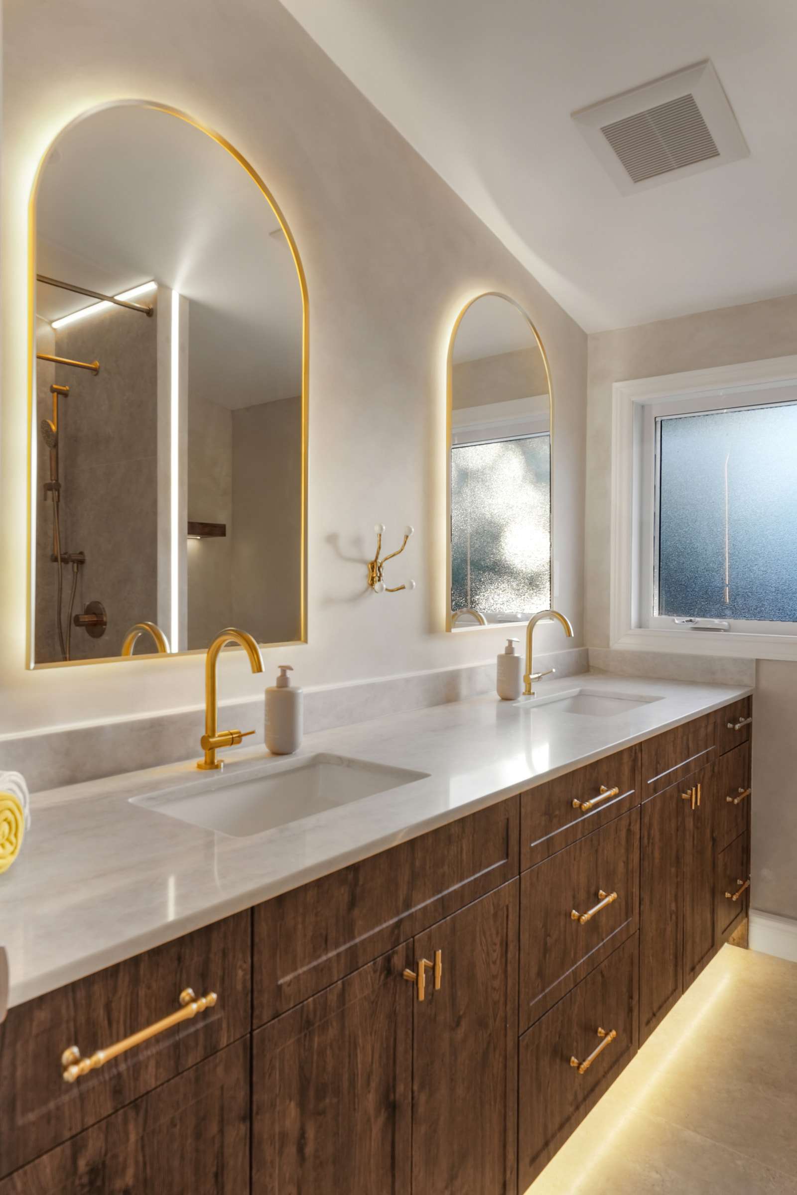 A double vanity under two backlit arched mirrors, lit from beneath, with a frosted window beside it.
