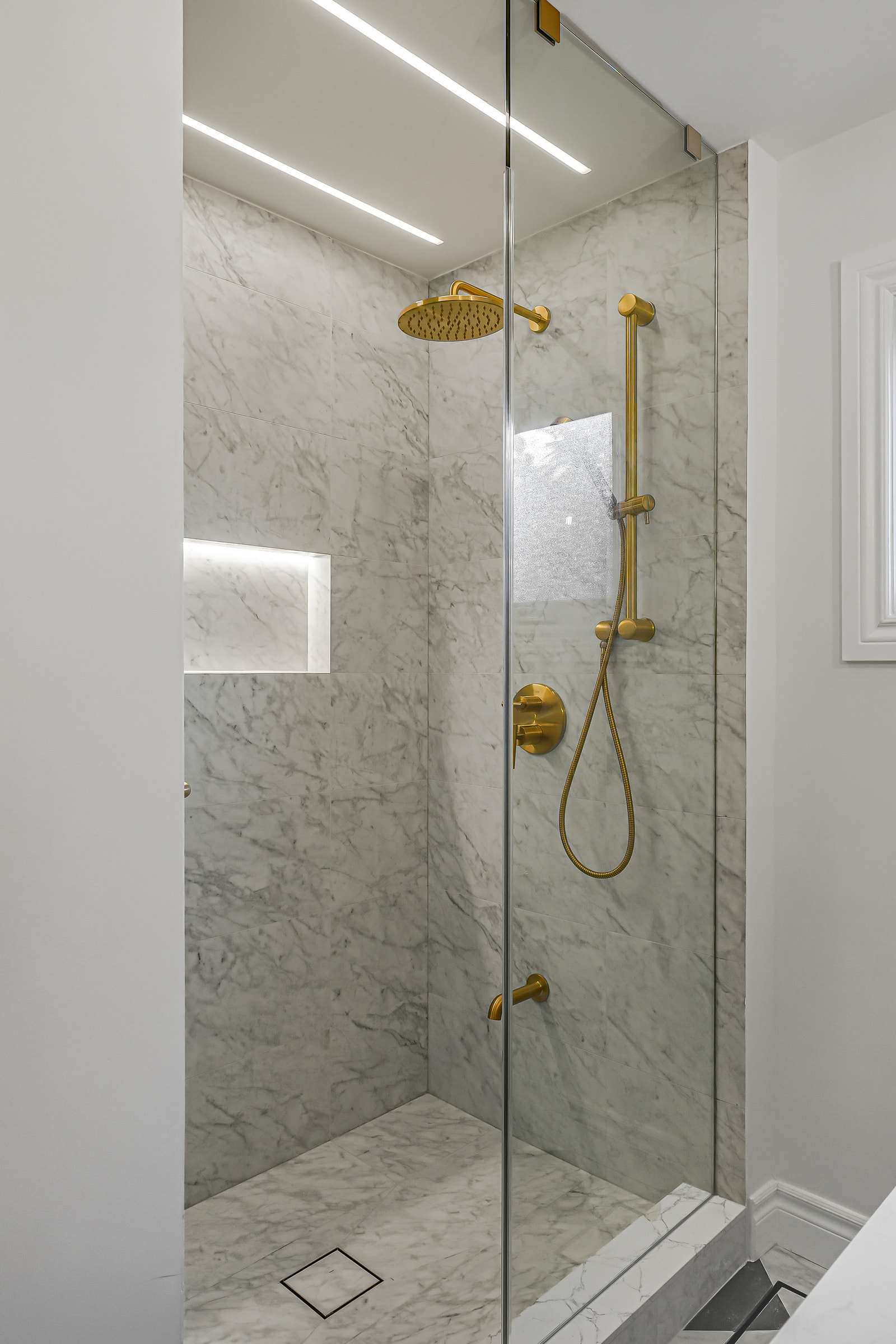 The walk-in shower behind clear glass, with a lit niche and two light lines in the ceiling.