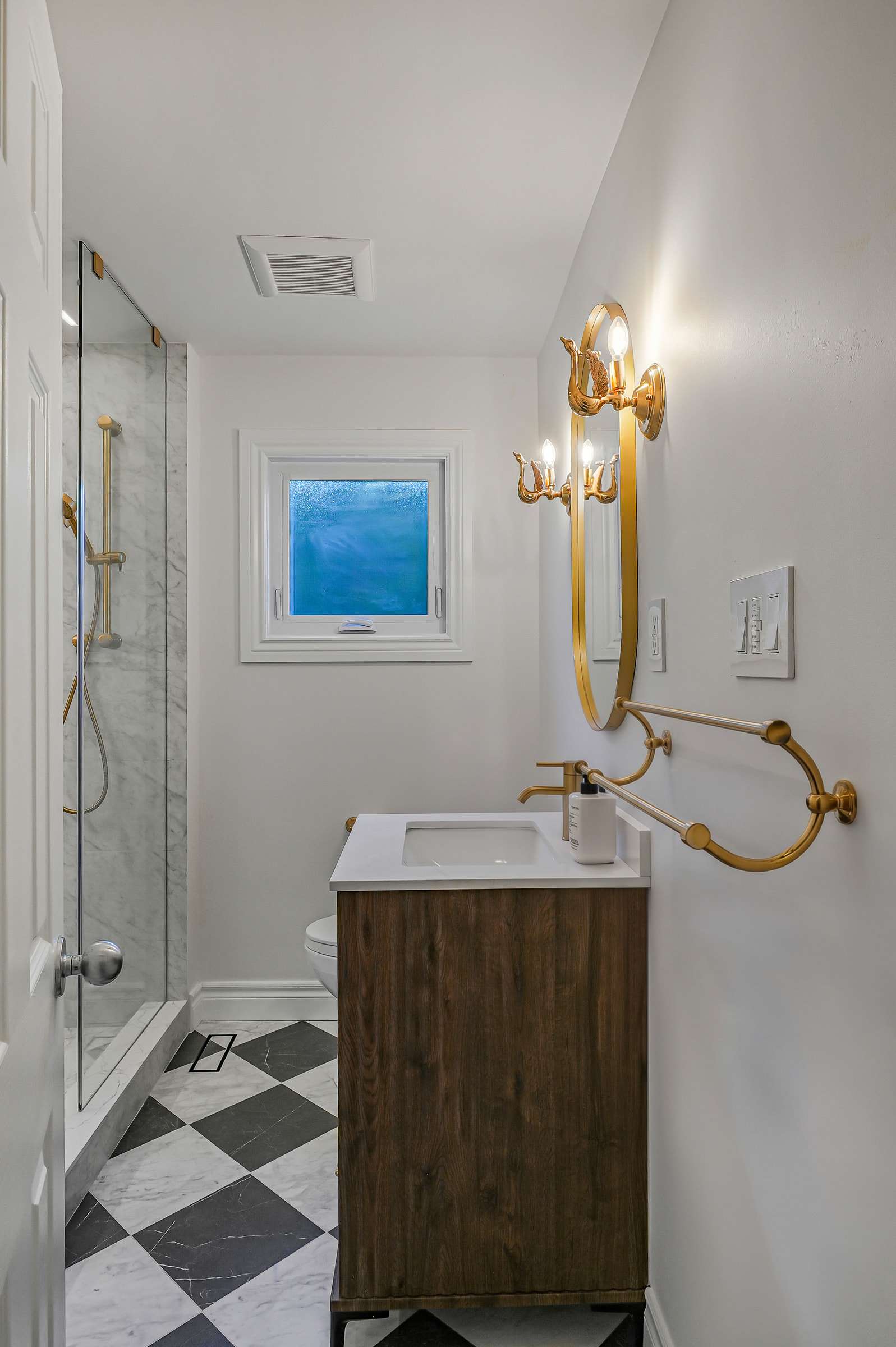 The shower room from the door: a glass walk-in shower, a vanity under an oval mirror between swan sconces, and a black-and-white floor.